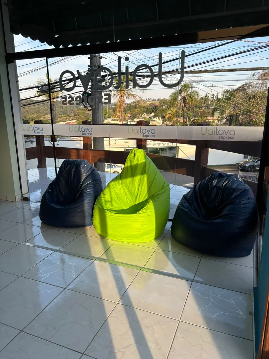 Área de espera da Uailava Express: três puffs, dois azuis e um verde-limão, virados para um janelão com vista das árvores e das casas de Bragança Paulista.