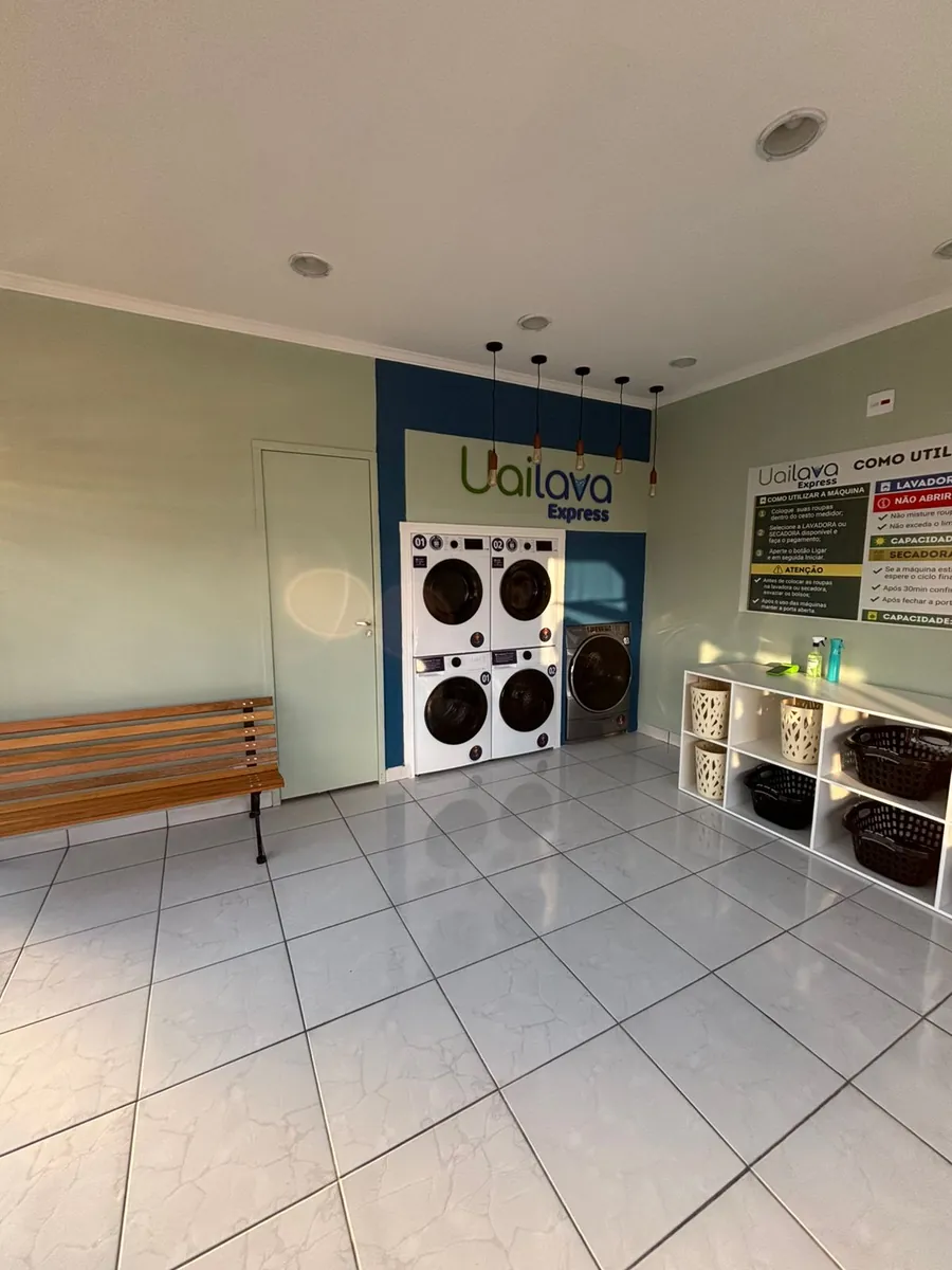 O salão visto da entrada, com um banco de madeira encostado na parede, as máquinas ao fundo sob o letreiro da Uailava e a estante de cestos à direita.